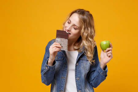 Young woman in denim clothes isolated on yellow orange background in studio. Proper nutrition healthy lifestyle sweets, dessert fast food, choice concept. Hold green apple eating biting chocolate bar.の写真素材