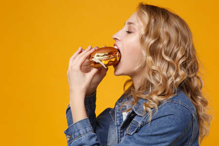Side view of young woman in denim clothes isolated on yellow orange wall background studio portrait. Proper nutrition or American classic fast food concept. Mock up copy space. Eating biting burger.の写真素材