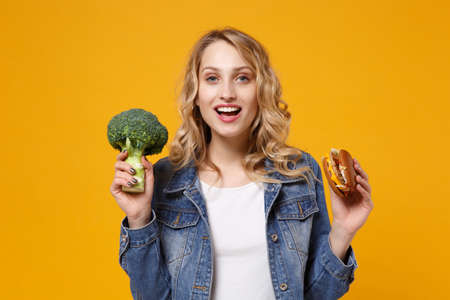 Cheerful young woman in denim clothes isolated on yellow orange wall background studio portrait. Proper nutrition, healthy lifestyle, fast food, choice concept. Holding in hands green broccoli burger.の写真素材