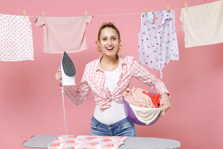 Excited young woman housewife in checkered shirt dry clothes on rope and ironing on board hold basin with clean clothes while doing housework isolated on pink background studio. Housekeeping concept.の写真素材
