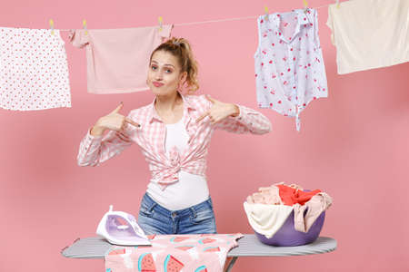 Young woman housewife in checkered shirt dry clothes on rope ironing clean clothes on board while doing housework isolated on pink background. Housekeeping concept. Pointing index fingers on herself.の写真素材