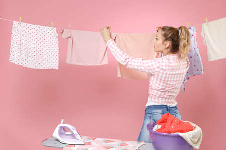 Beautiful young girl housewife in checkered shirt dry clothes on rope ironing clean clothes while doing housework isolated on pink background studio portrait. Housekeeping concept. Mock up copy space.の写真素材