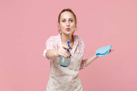 Cheerful young woman housewife in apron hold atomizer spray with washing cleanser, cleaning rag...