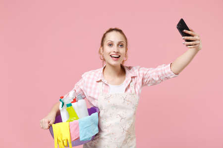Excited young woman housewife in apron hold basin with detergent bottles washing cleansers while doing housework isolated on pink background. Housekeeping concept. Doing selfie shot on mobile phone.の写真素材