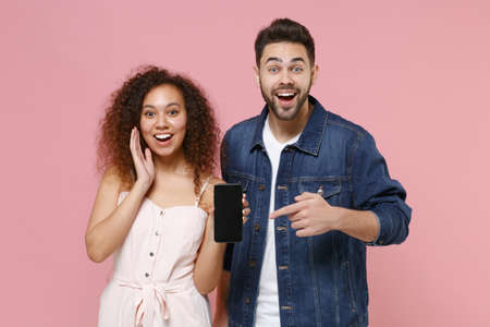 Amazed young couple friends european guy african american girl in casual clothes isolated on pink background. People lifestyle concept. Pointing index finger on mobile phone with blank empty screen.の写真素材