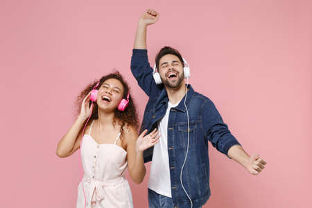 Funny young couple friends european guy african american girl in casual clothes isolated on pastel pink background. People lifestyle concept. Listen music with headphones keeping eyes closed, dancing.の写真素材