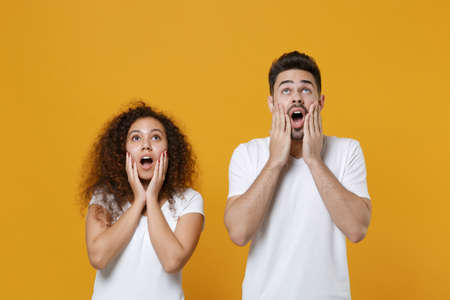 Shocked young couple friends european guy african american girl in white t-shirts posing isolated on yellow background. People lifestyle concept. Put hands on cheeks, looking up, keeping mouth open.の写真素材