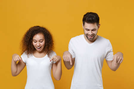 Amazed young couple two friends european guy african american girl in white t-shirts isolated on yellow background in studio. People lifestyle concept. Mock up copy space. Pointing index fingers down.の写真素材