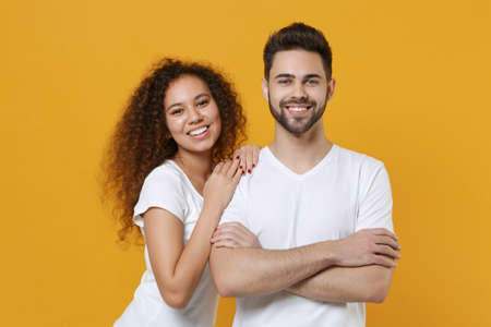 Smiling young couple two friends european guy african american girl in white t-shirts posing isolated on yellow background studio. People lifestyle concept. Mock up copy space. Holding hands crossed.の写真素材