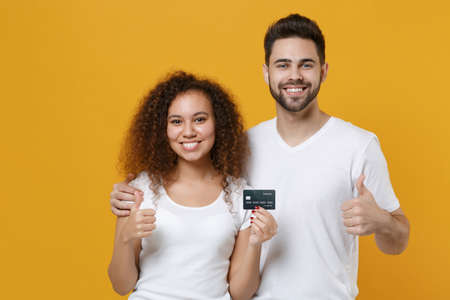 Smiling young couple two friends european guy african american girl in white t-shirts posing isolated on yellow background studio. People lifestyle concept. Hold credit bank card, showing thumbs up.の写真素材
