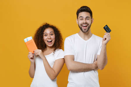 Excited young couple two friends european guy african american girl in white t-shirts posing isolated on yellow background in studio. People lifestyle concept. Hold credit bank card, passport tickets.