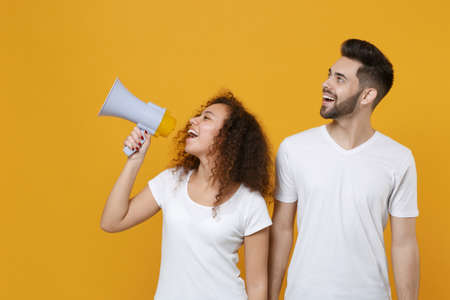 Cheerful couple two friends european guy african american girl in white t-shirts posing isolated on yellow background. People lifestyle concept. Mock up copy space. Scream in megaphone, looking aside.の写真素材