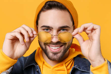 Close up of smiling young hipster guy in fashion jeans denim clothes posing isolated on yellow orange wall background studio portrait. People lifestyle concept. Mock up copy space. Holding glasses.の写真素材