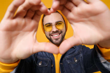 Smiling young hipster guy in fashion jeans denim clothes posing isolated on yellow orange background. People lifestyle concept. Mock up copy space. Showing shape heart with hands, heart-shape sign.の写真素材