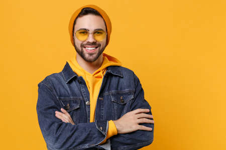 Smiling young hipster guy in fashion jeans denim clothes posing isolated on yellow orange background studio portrait. People emotions lifestyle concept. Mock up copy space. Holding hands crossed.の写真素材