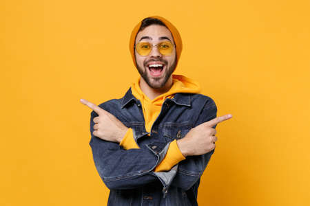 Excited young hipster guy in fashion jeans denim clothes posing isolated on yellow orange background. People lifestyle concept. Mock up copy space. Pointing index fingers aside, holding hands crossed.の写真素材