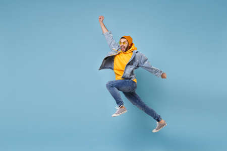 Crazy young hipster guy in fashion jeans denim clothes posing isolated on pastel blue background. People lifestyle concept. Mock up copy space. Jumping, spreading hands, clenching fists like winner.の写真素材