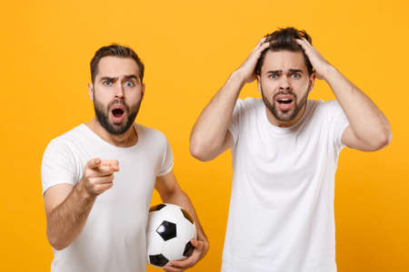 Shocked men guys friends in white t-shirt posing isolated on yellow background. Sport leisure concept. Mock up copy space. Cheer up support favorite team with soccer ball point index finger on camera.の写真素材