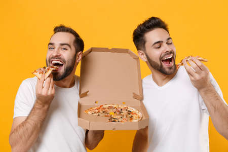 Close up of funny young men guys friends in white blank t-shirts posing isolated on yellow orange background. People lifestyle concept. Mock up copy space. Holding italian pizza in cardboard flatbox.の写真素材