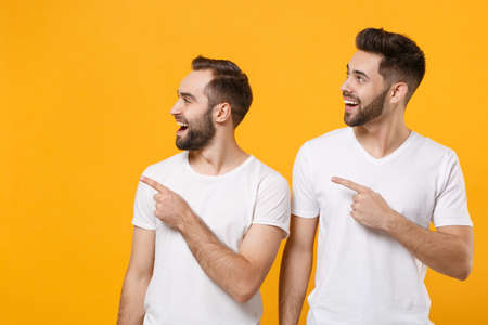 Cheerful young men guys friends in white blank empty t-shirts posing isolated on yellow orange background studio portrait. People lifestyle concept. Mock up copy space. Pointing index fingers aside.の写真素材