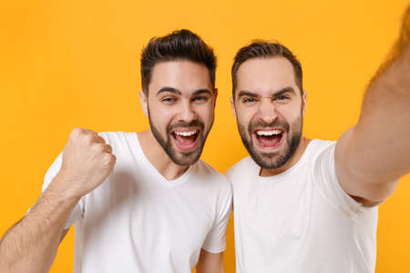 Close up of joyful young men guys friends in white t-shirts isolated on yellow orange background....
