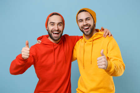 Two cheerful hipster men guys in fashion red yellow clothes posing isolated on pastel blue background studio portrait. People sincere emotions lifestyle concept. Mock up copy space. Showing thumbs up.の写真素材