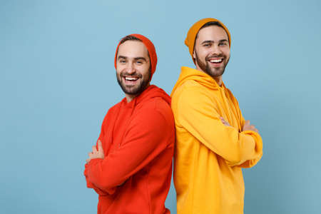 Two cheerful hipster men guys in fashion red yellow clothes posing isolated on pastel blue background studio portrait. People emotions lifestyle concept. Mock up copy space. Holding hands crossed.の写真素材