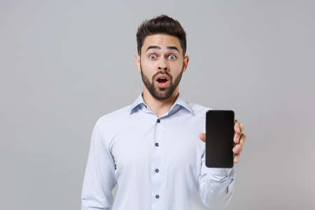 Shocked young unshaven business man in light shirt posing isolated on grey wall background. Achievement career wealth business concept. Mock up copy space. Hold mobile phone with blank empty screen.の写真素材
