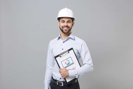 Smiling young unshaven business man in light shirt, protective construction white helmet isolated on...