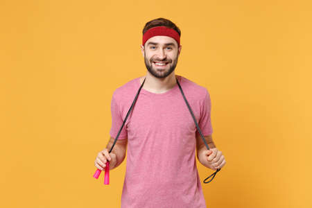 Smiling young bearded fitness sporty guy 20s sportsman in headband t-shirt spend weekend in home gym isolated on yellow background. Workout sport motivation lifestyle concept. Hold skipping rope.の写真素材