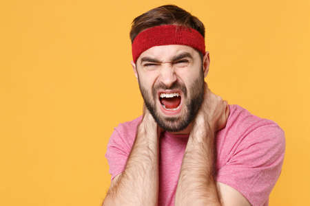 Screaming bearded fitness sporty guy 20s sportsman in headband t-shirt spend weekend in home gym isolated on yellow background. Workout sport motivation lifestyle concept. Touching neck, get injuredの写真素材