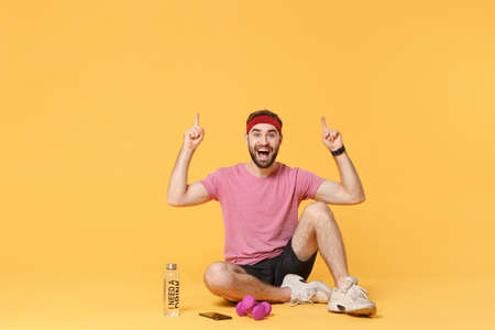Excited fitness guy sportsman in headband t-shirt in home gym isolated on yellow background. Workout sport motivation concept. Sit with smart watch, water mobile phone dumbbell point index fingers up.の写真素材