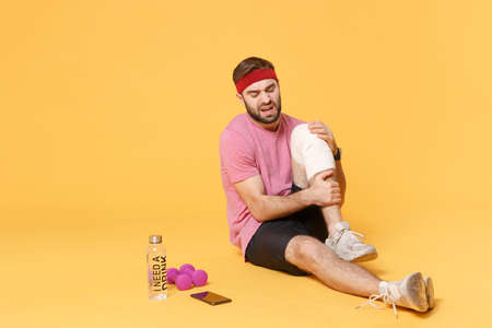 Ingured displeased guy sportsman in t-shirt in home gym isolated on yellow background. Workout sport motivation concept. Sit with water bottle mobile phone dumbbells touch knee with elastic bandage.の写真素材