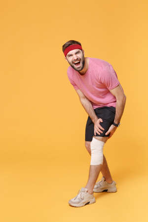 Ingured screaming young bearded fitness guy 20s sportsman in headband t-shirt in home gym isolated on yellow background. Workout sport motivation lifestyle concept. Touch knee with elastic bandage.の写真素材