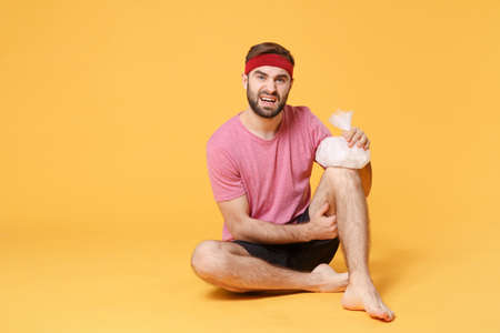Displeased young bearded fitness sporty guy sportsman in headband t-shirt in home gym isolated on yellow background. Workout sport motivation concept. Get injured hold ice pack cooling down knee pain.の写真素材