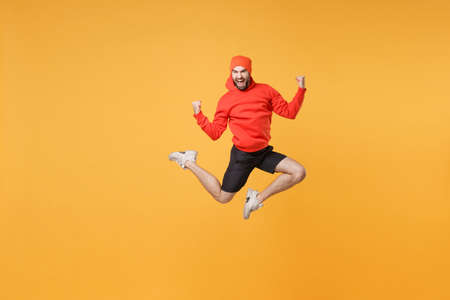 Excited young bearded fitness sporty guy 20s sportsman in hat hoodie shorts in home gym isolated on yellow background. Workout sport motivation lifestyle concept. Jump doing winner gesture screaming.の写真素材