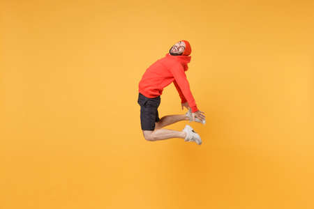 Side view of cheerful young fitness sporty guy sportsman in hat hoodie shorts in home gym isolated on yellow background. Workout sport motivation lifestyle concept. Jumping spreading hands, screaming.の写真素材
