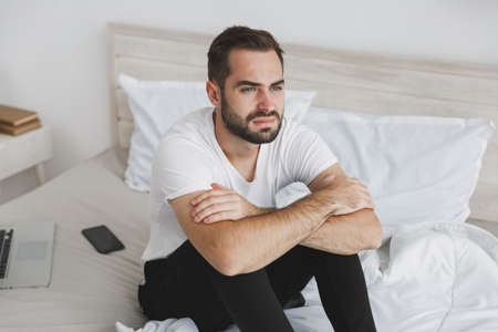 Calm young handsome bearded man sitting in bed with white sheet pillow blanket in bedroom at home. Sad upset male spending time in room. Rest relax good mood lifestyle concept. Mock up copy space.の写真素材