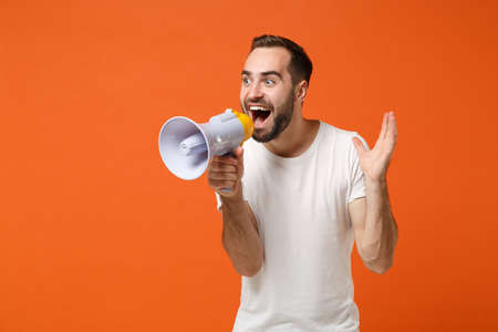 Excited young man in casual white t-shirt posing isolated on bright orange wall background studio portrait. People sincere emotions lifestyle concept. Mock up copy space. Screaming in megaphone.の写真素材