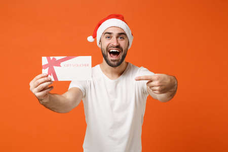 Excited young Santa man in white t-shirt, Christmas hat posing isolated on orange background. New Year 2020 celebration holiday concept. Mock up copy space. Pointing index finger on gift certificate.の写真素材