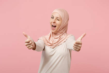 Cheerful young arabian muslim woman in hijab light clothes posing isolated on pink background studio...