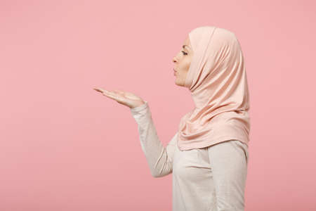 Side view of arabian muslim girl in hijab light clothes posing isolated on pink wall background studio portrait. People religious Islam lifestyle concept. Mock up copy space. Blowing sending air kiss.の写真素材