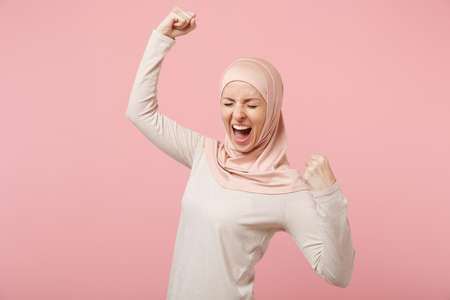 Happy young arabian muslim woman in hijab light clothes posing isolated on pink background. People religious Islam lifestyle concept. Mock up copy space. Screaming, doing winner gesture, saying Yes.の写真素材