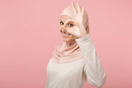 Arabian muslim woman in hijab light clothes posing isolated on pink background. People religious lifestyle concept. Mock up copy space. Showing OK gesture, holding hand near eye, imitating binoculars.の写真素材