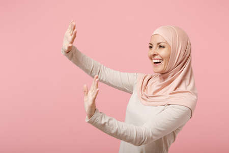 Cheerful young arabian muslim woman in hijab light clothes posing isolated on pink background....