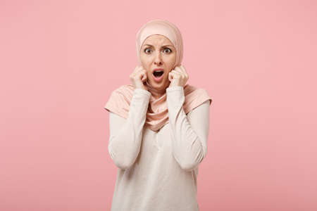 Shocked scared arabian muslim woman in hijab light clothes posing isolated on pink background. People religious Islam lifestyle concept. Mock up copy space. Keeping mouth open putting hands on cheeks.の写真素材