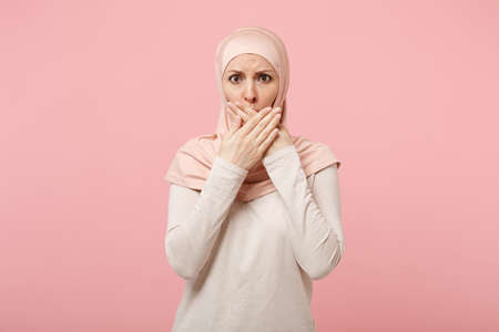 Disturbed young arabian muslim woman in hijab light clothes posing isolated on pink background studio portrait. People religious Islam lifestyle concept. Mock up copy space. Covering mouth with hands.の写真素材