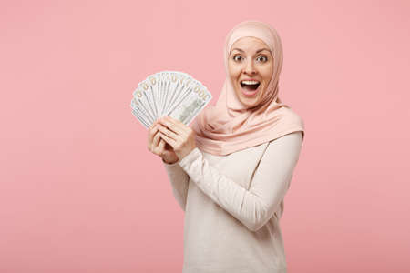 Excited young arabian muslim woman in hijab light clothes posing isolated on pink background. People religious Islam lifestyle concept. Mock up copy space. Hold fan of cash money in dollar banknotes.の写真素材
