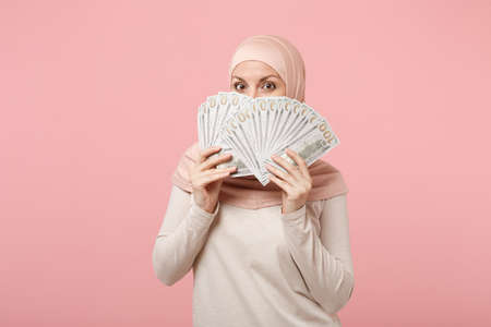 Arabian muslim woman in hijab light clothes posing isolated on pink background. People religious Islam lifestyle concept. Mock up copy space. Covering face with fan of cash money in dollar banknotes.の写真素材