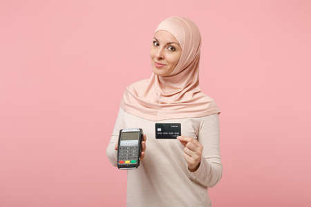 Arabian muslim woman in hijab light clothes posing isolated on pink background. People religious lifestyle concept. Mock up copy space. Hold payment terminal to process, acquire credit card payments.の写真素材